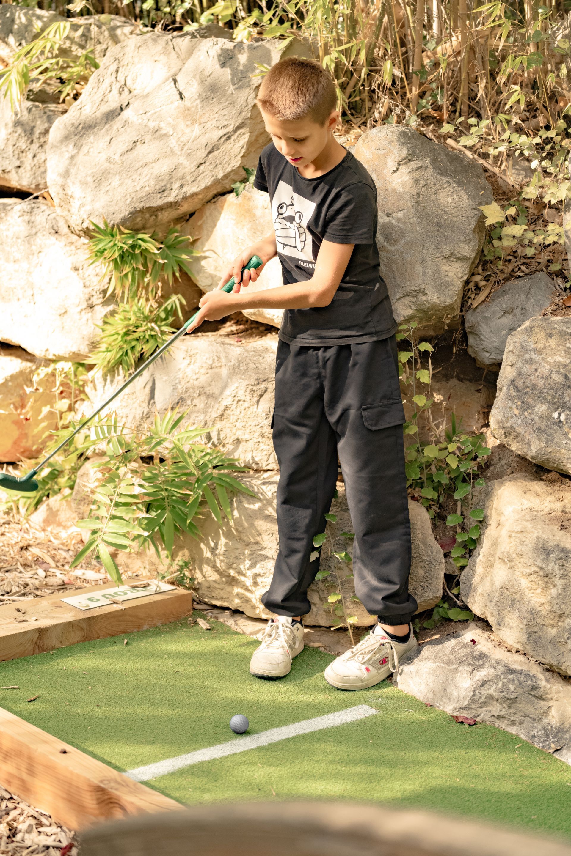 Garçon vêtu de noir jouant au mini-golf, visant une balle avec son club sur un terrain gazonné et rocheux en arrière-plan.