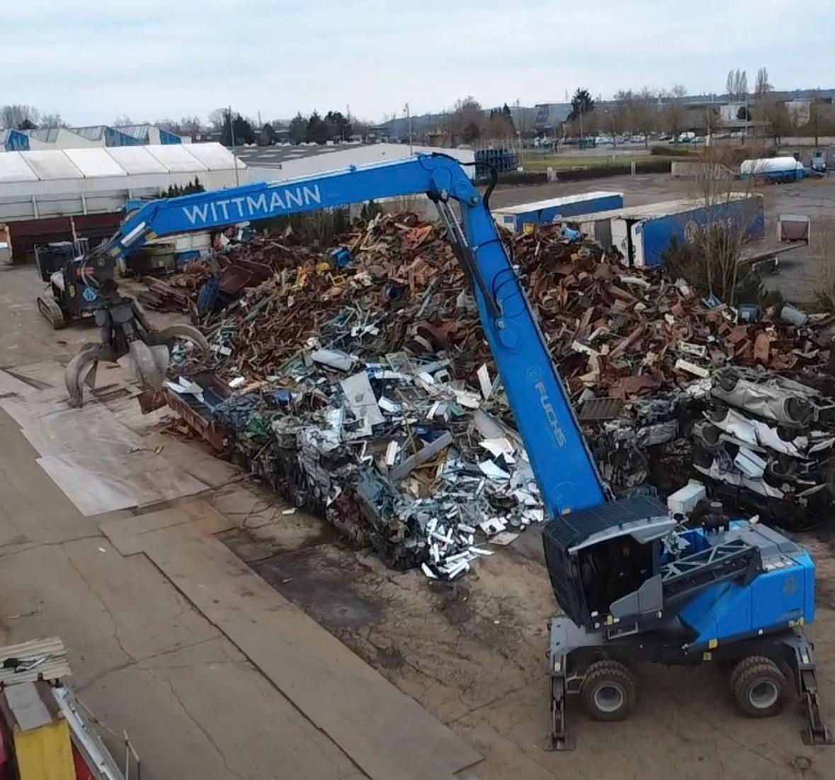 Vue aérienne d'un véhicule avec grue et pince devant un tas de métaux.