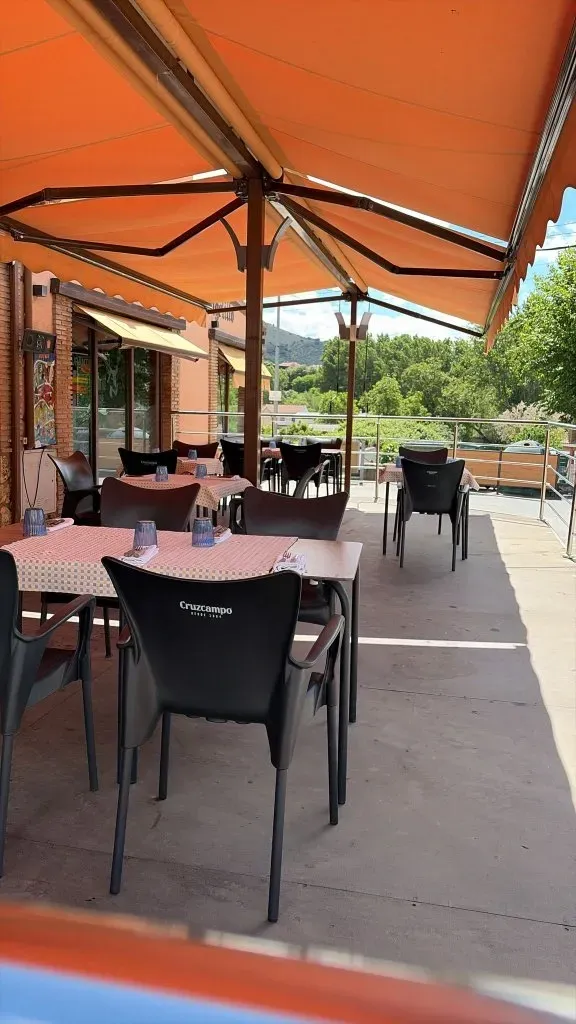 Terraza de un restaurante al aire libre con toldo naranja, sillas negras y mesas en un día soleado.