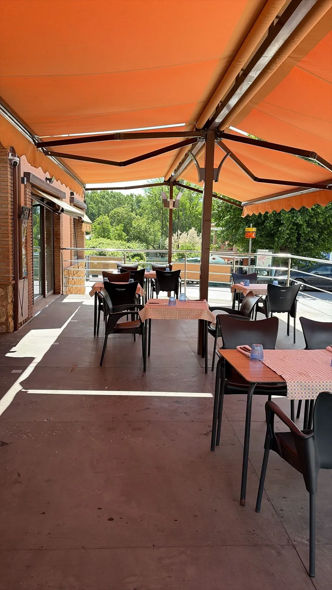 Terraza cubierta de una cafetería con toldo naranja, mesas vacías y coches aparcados al fondo.