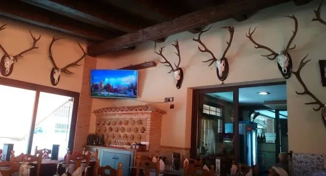 Interior acogedor y rústico del restaurante con astas de ciervo montadas, un televisor en la pared y mesas de comedor cerca de una puerta con ventana.