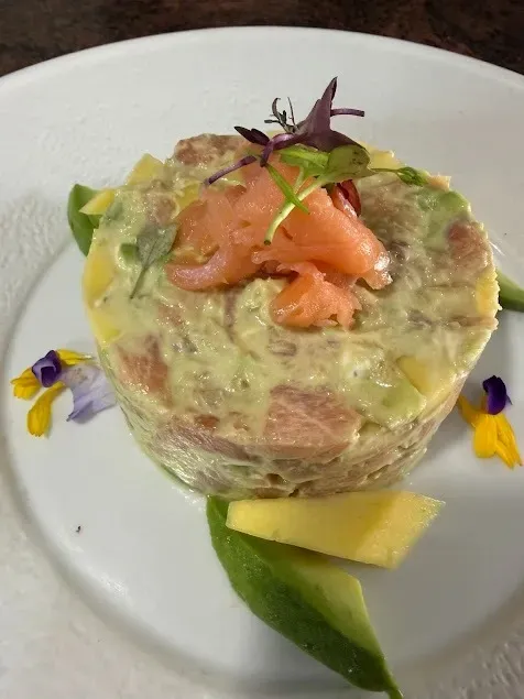 Tartar de salmón en un molde de cristal con forma de anillo, decorado con hierbas aromáticas, sobre un plato blanco con rodajas de aguacate y mango.
