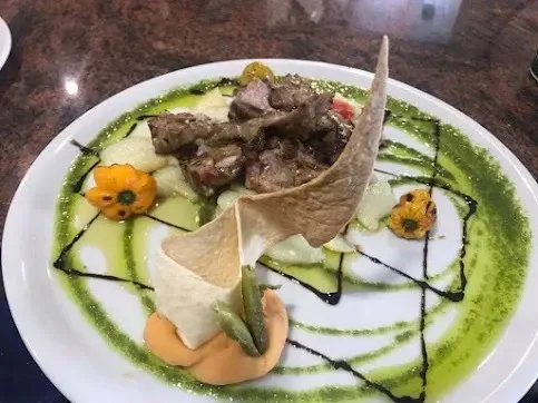 Plato gourmet de carne emplatado con un chorrito de salsa verde, una teja decorativa y pequeñas guarniciones de verduras sobre un plato blanco.