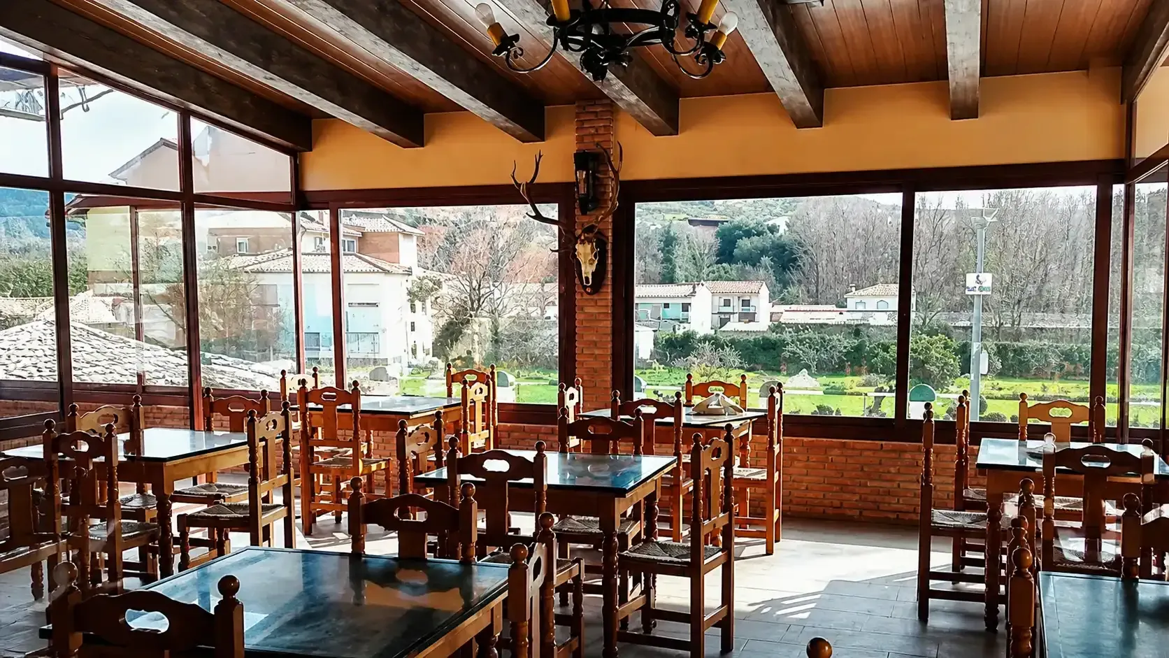 Cafetería rústica vacía con mesas y sillas de madera, grandes ventanales y vistas a la vegetación exterior.