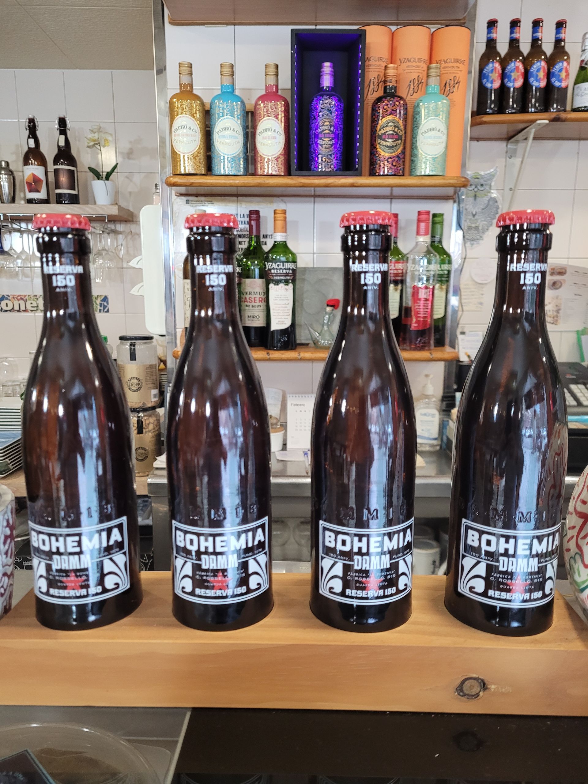 Cuatro grandes botellas de cerveza Bohemia sobre un mostrador de madera, con botellas de licor en estantes al fondo.