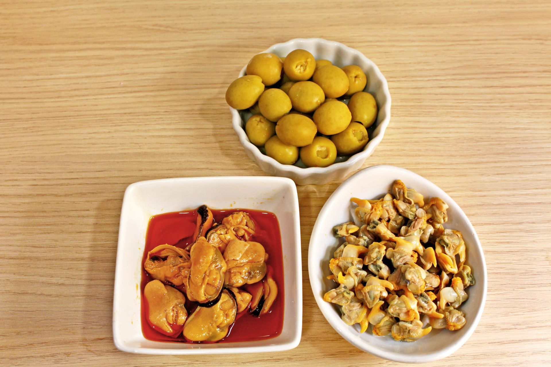 Tres pequeños cuencos de comida: aceitunas, mejillones y almejas sobre una mesa de madera.