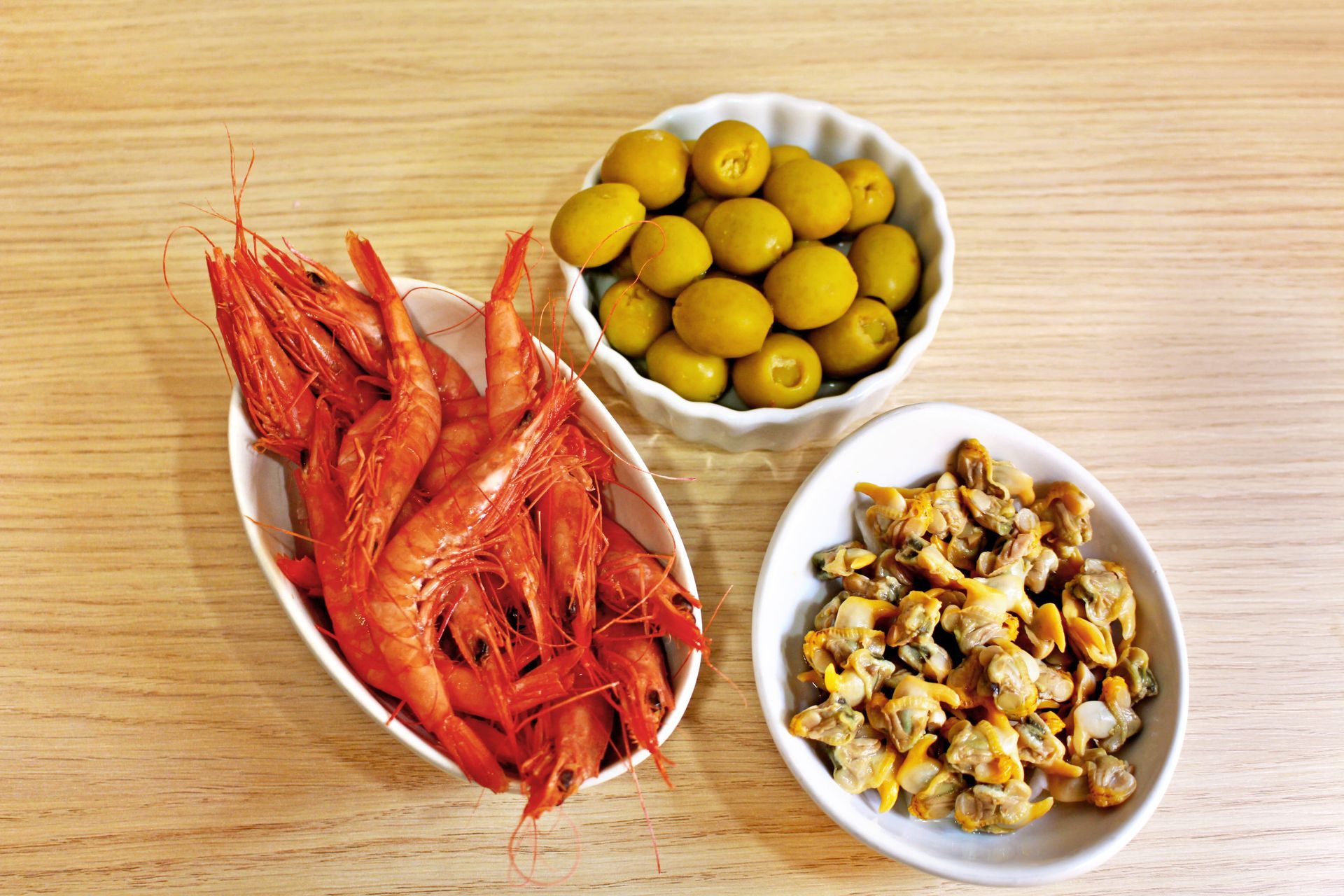 Camarones, aceitunas y almejas en platos blancos sobre una mesa de madera.
