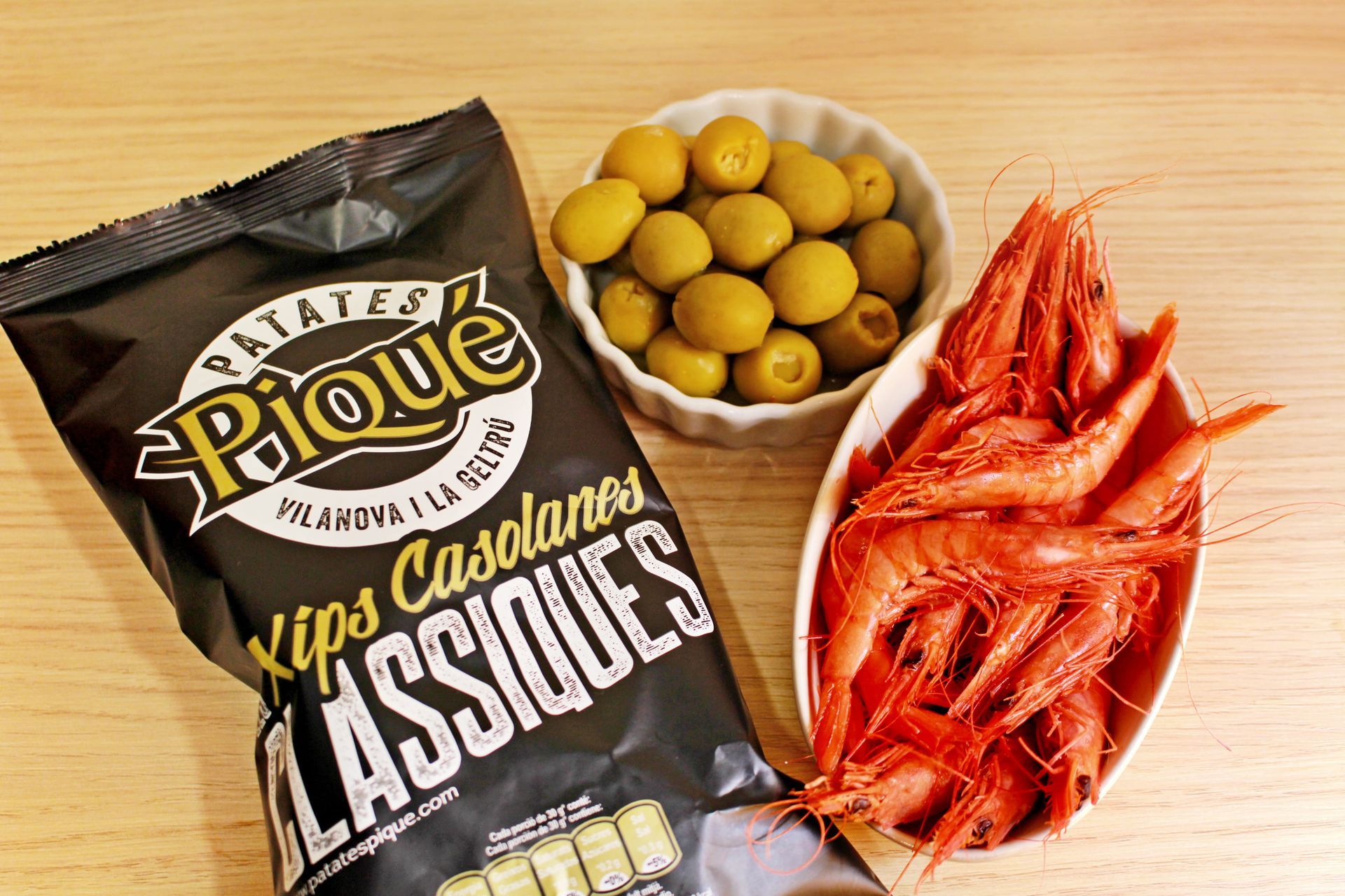 Una bolsa de patatas fritas, un bol de aceitunas y un bol de gambas rojas sobre una superficie de madera.