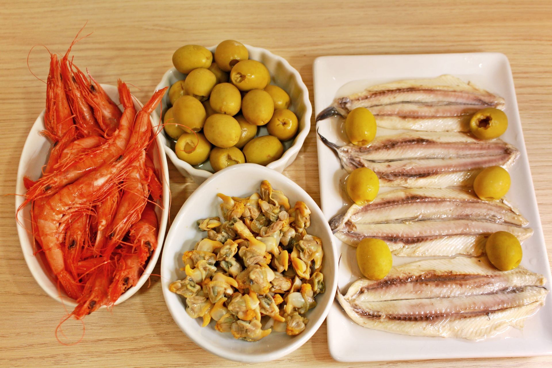 Tapas de marisco: gambas, aceitunas, berberechos y anchoas sobre mesa de madera.