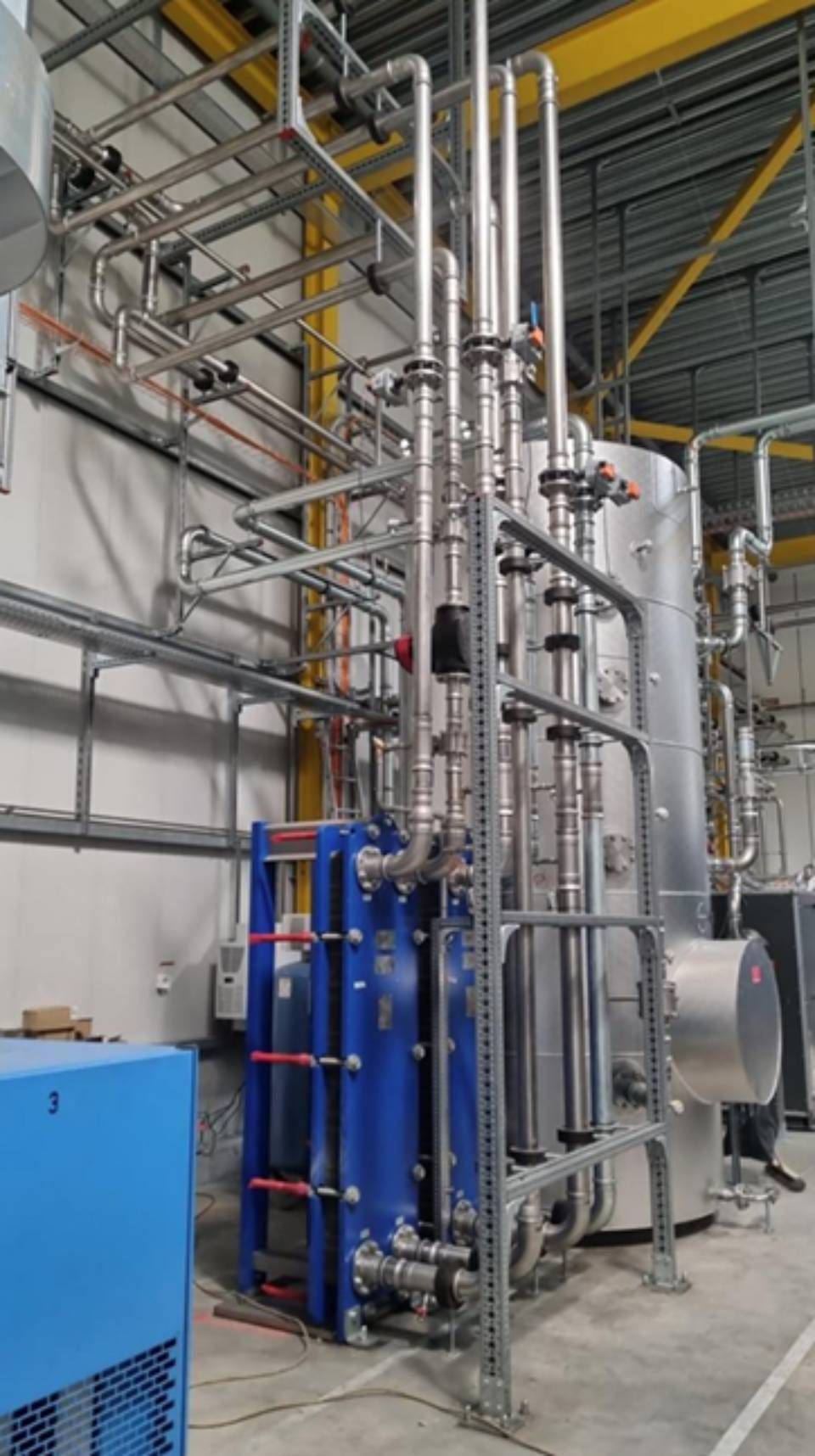 Industriemaschinen mit blauen und silbernen Bauteilen, umgeben von Rohren und in einer Fabrikumgebung angeordnet.