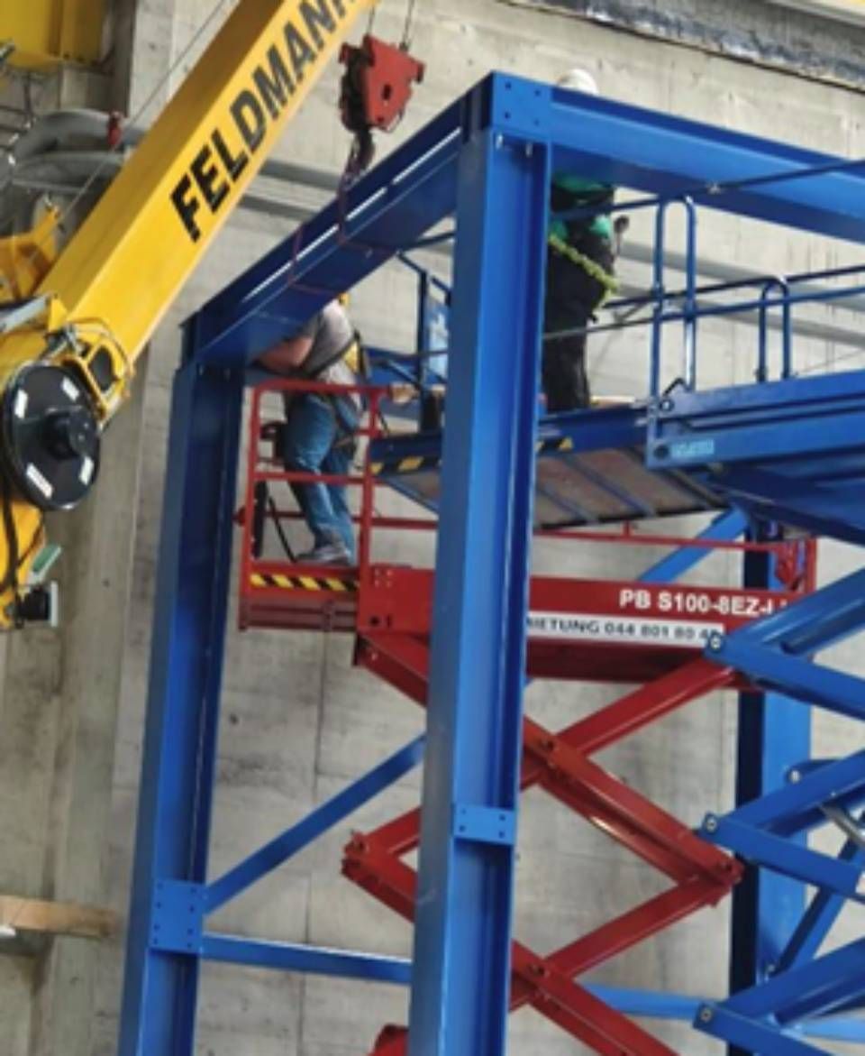 Arbeiter montieren mit einem Hubwagen einen blauen Stahlrahmen; Kran hebt ein Teilstück an.