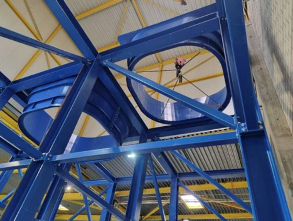 Ein blaues Metallgerüst stützt gebogene blaue Leitungen. In einem dieser Rohre arbeitet eine Person.