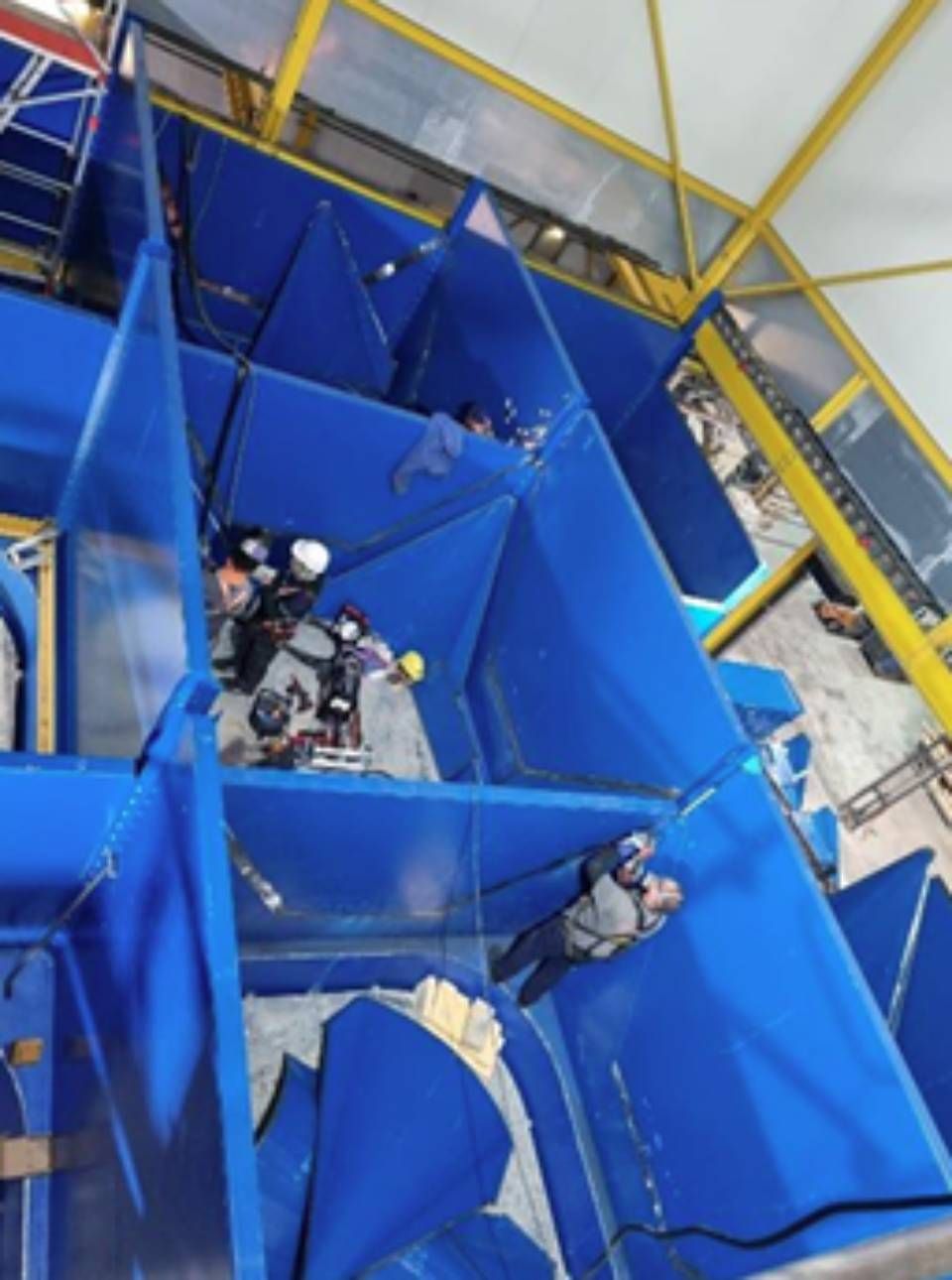 Arbeiter in einer grossen blauen Industrieanlage, möglicherweise einer Rutsche oder einem Trichter. Sie tragen Schutzhelme, einige auch Atemschutzmasken.