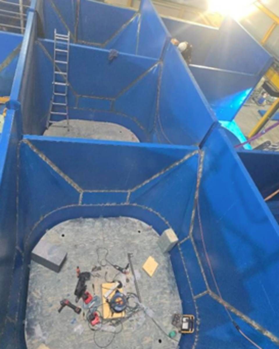 Blaue, im Bau befindliche Metallkonstruktion mit einer Leiter und Werkzeugen im Inneren; Arbeiter sind zu sehen.