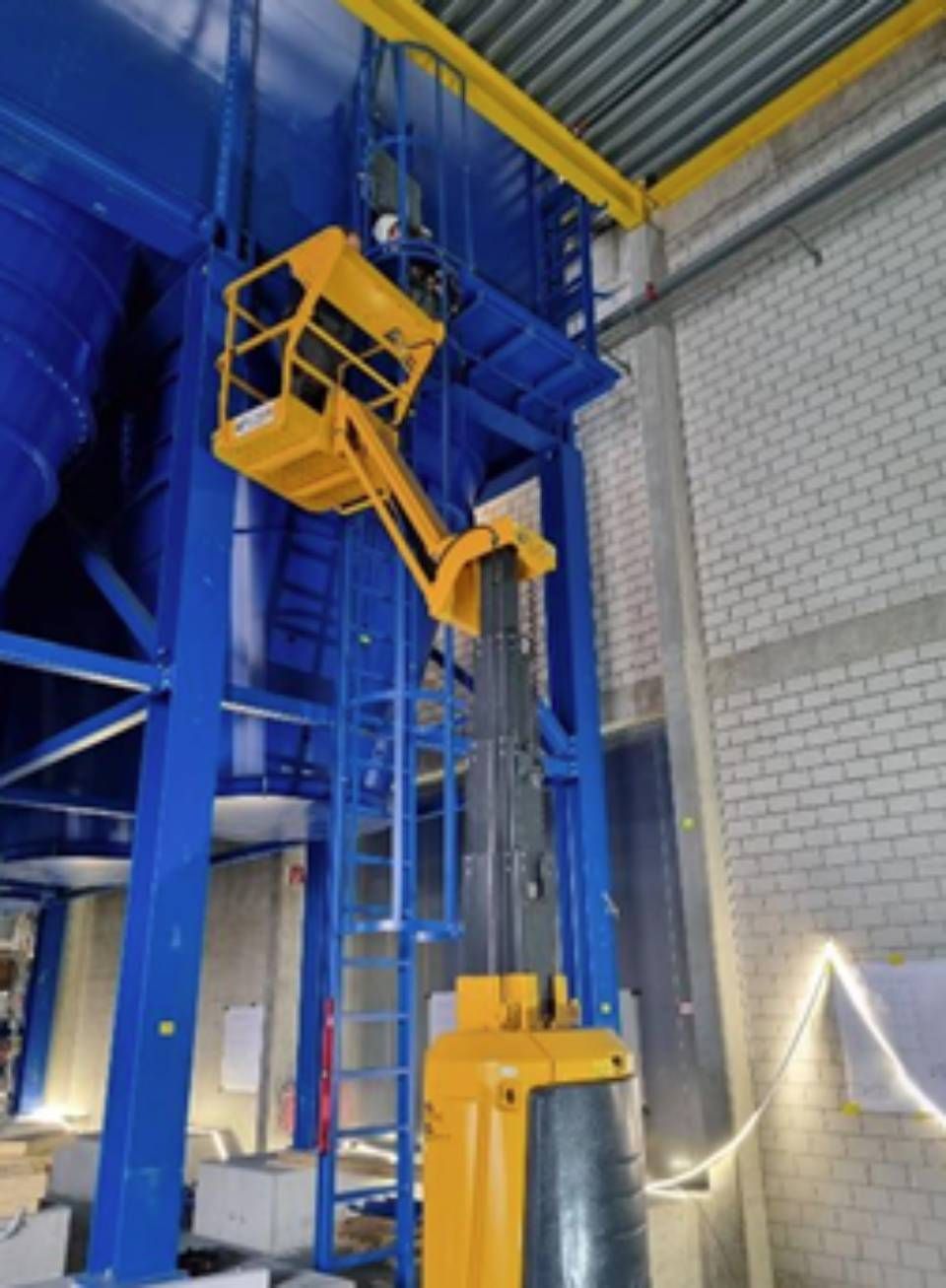 Gelber Aufzug mit einem Arbeiter darin, der nach einer blauen Industrieanlage im Inneren greift.