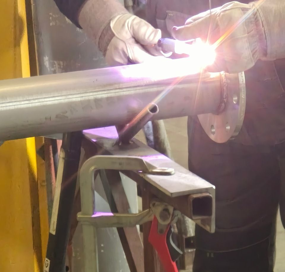 Ein Schweisser verbindet in einer Werkstatt mit einem Schweissbrenner Metallrohre; helle Funken und Schutzhandschuhe sind sichtbar.