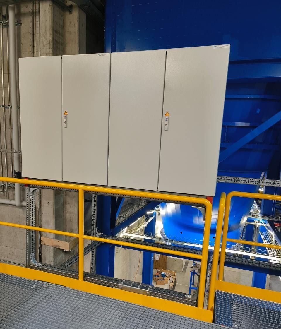 Weisse Schalttafeln sind an einem gelben Geländer montiert und bieten einen Blick auf die Maschinen in einer Fabrikanlage.