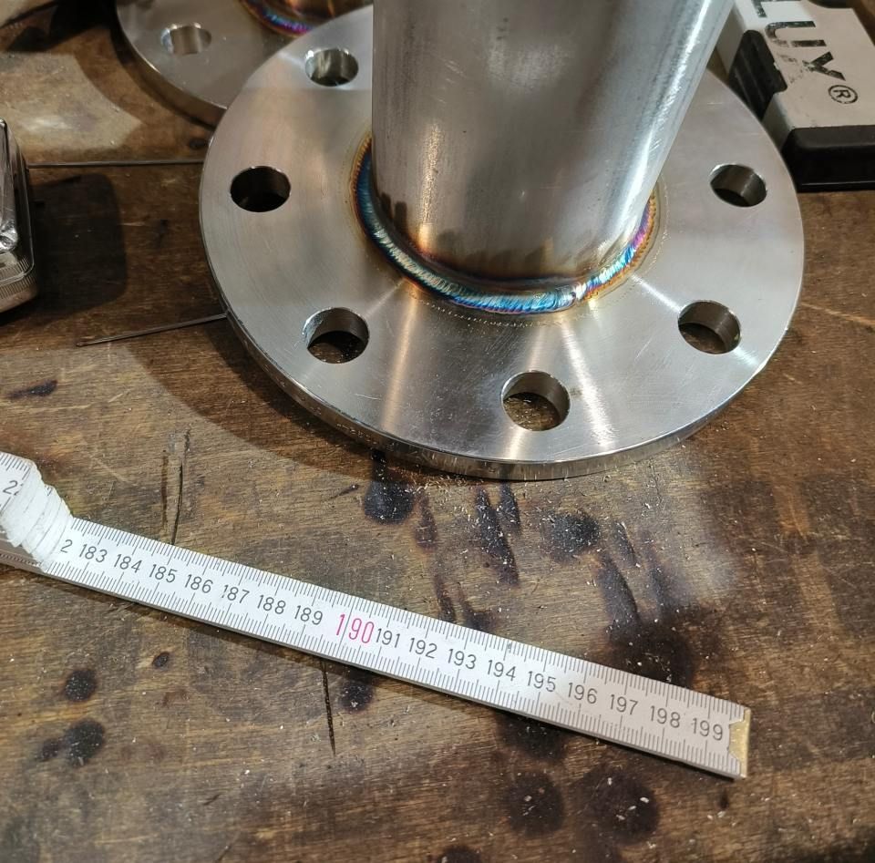 Ein an einem Flansch auf einer Holzoberfläche angeschweißtes Metallrohr, daneben ein Lineal.