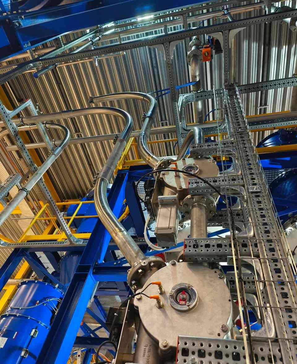 Komplexe Industrieanlagen mit Rohren und Metallkonstruktionen in einem Raum mit hoher Decke. Blaue und gelbe Akzente.