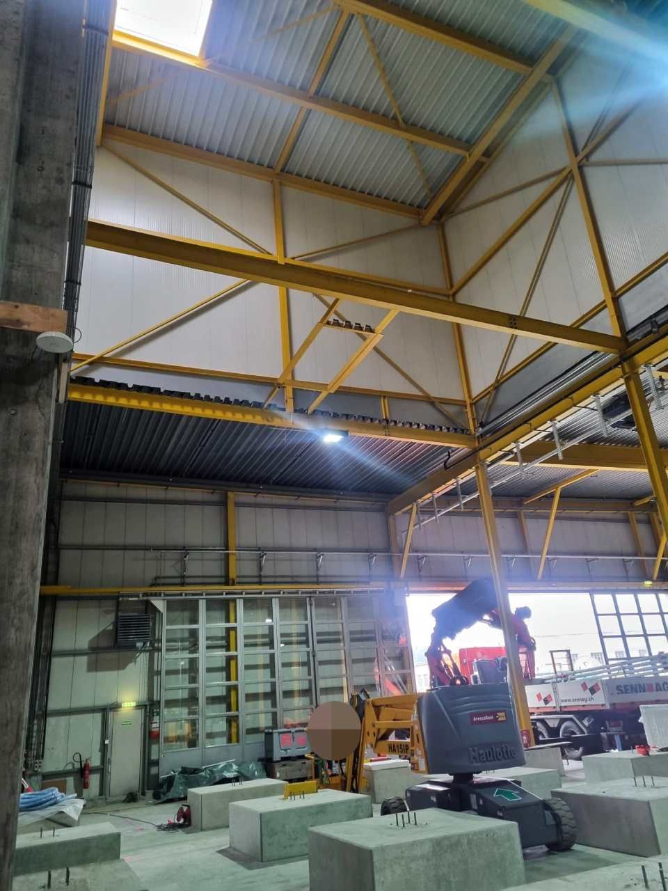 Innenansicht eines im Bau befindlichen Industriegebäudes: gelbe Metallträger, Betonblöcke, Baumaschinen.