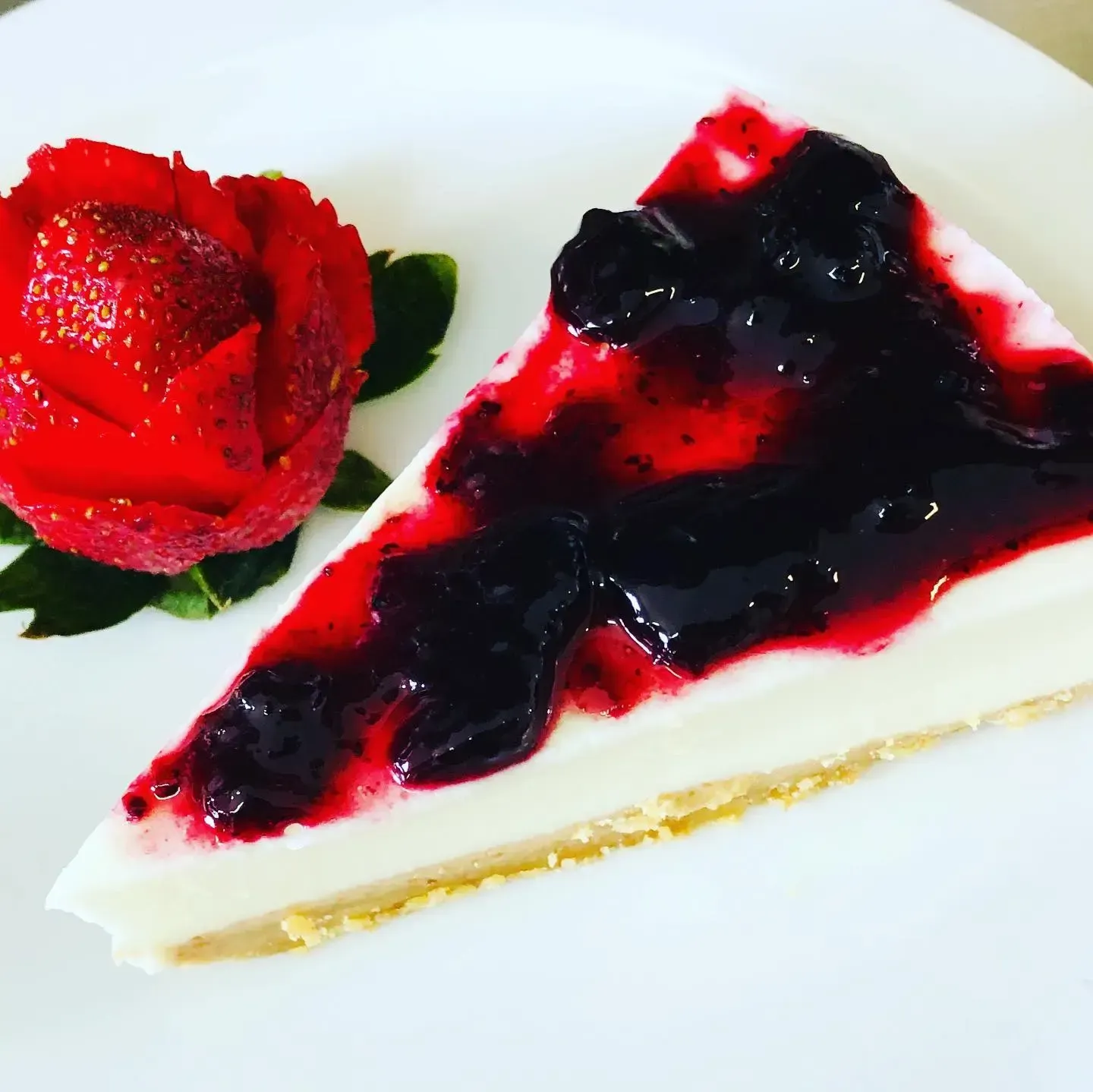Una porción de tarta de queso con cobertura de frutos rojos oscuros junto a una rosa de fresa roja.
