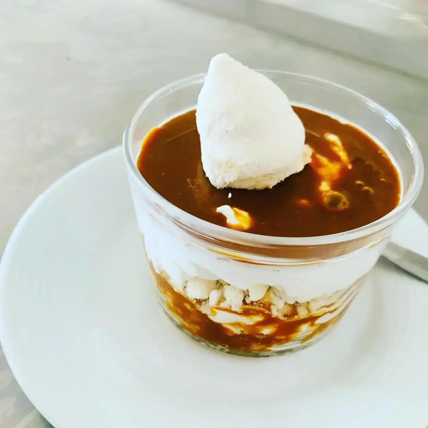 Postre por capas en un vaso transparente, coronado con una bola de helado blanca, sobre un plato blanco.