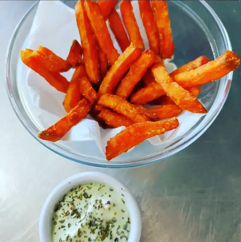 Un bol de patatas fritas crujientes de color naranja con una pequeña taza de salsa de hierbas para mojar sobre una mesa de metal.