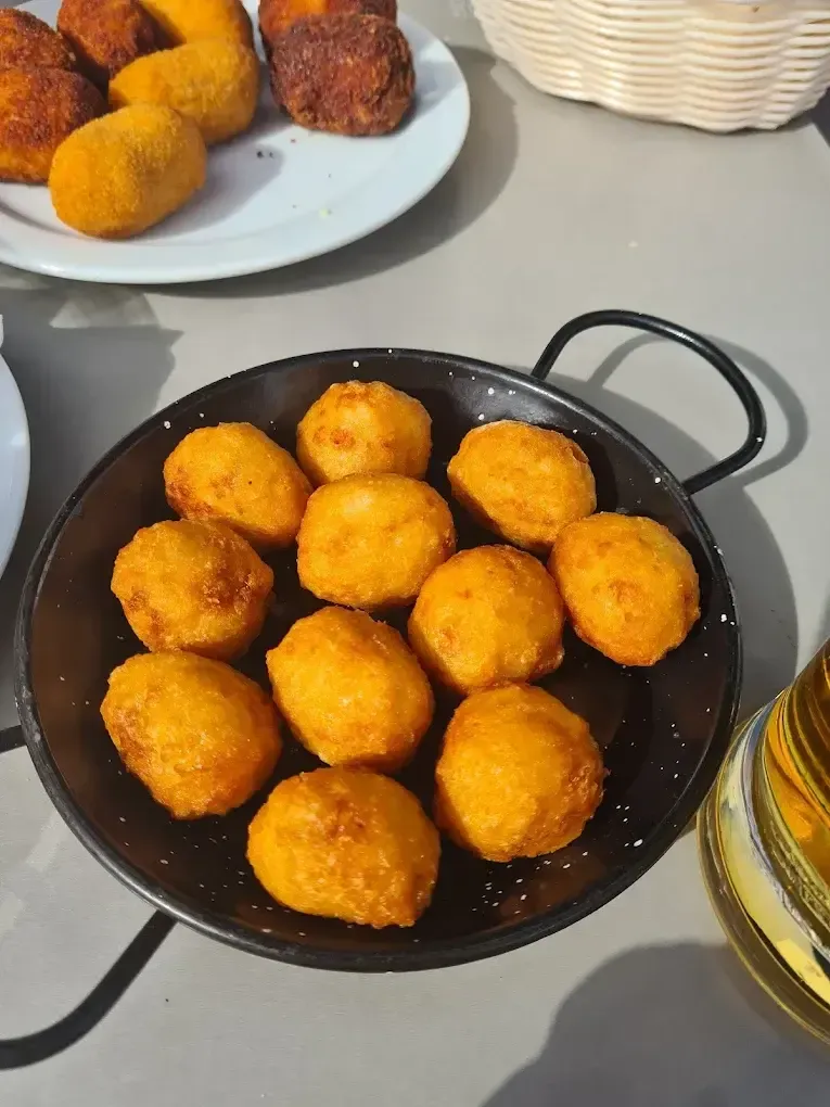 Una pequeña sartén negra llena de croquetas fritas doradas sobre una mesa gris, con un plato y un vaso cerca.