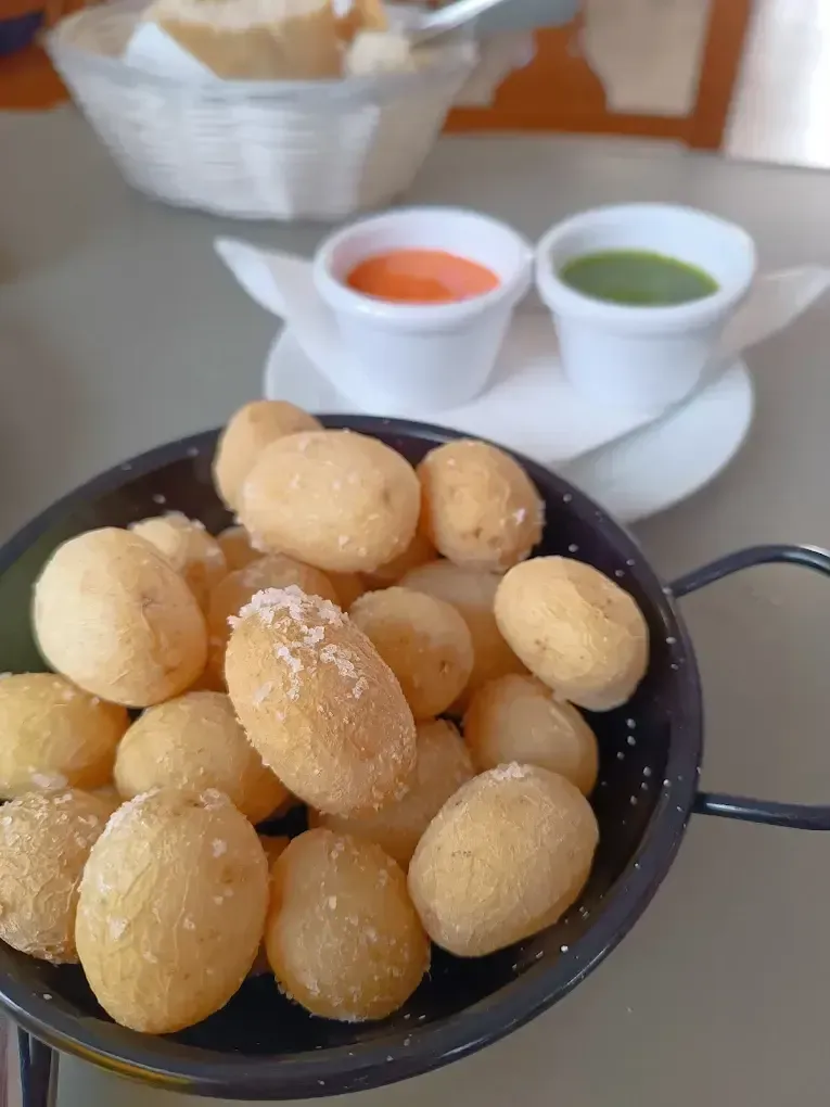 Bolitas de masa frita en un colador negro, con salsas para mojar de color naranja y verde sobre una mesa blanca.