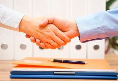 Dos personas se dan la mano sobre una pila de archivos, con un bolígrafo encima. Entorno de oficina.