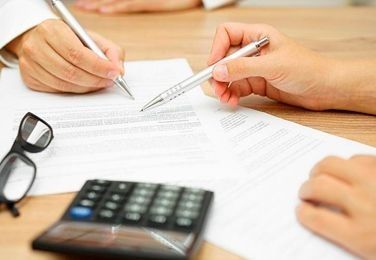 Manos firmando documentos con bolígrafos; calculadora y gafas sobre el escritorio.