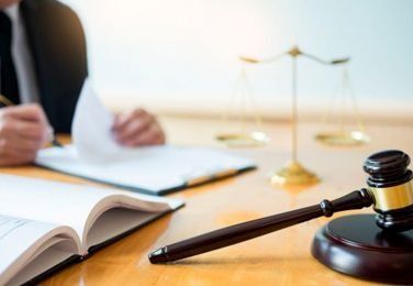 Mazo sobre un escritorio de madera con una balanza de la justicia, un libro de leyes y una persona revisando documentos.