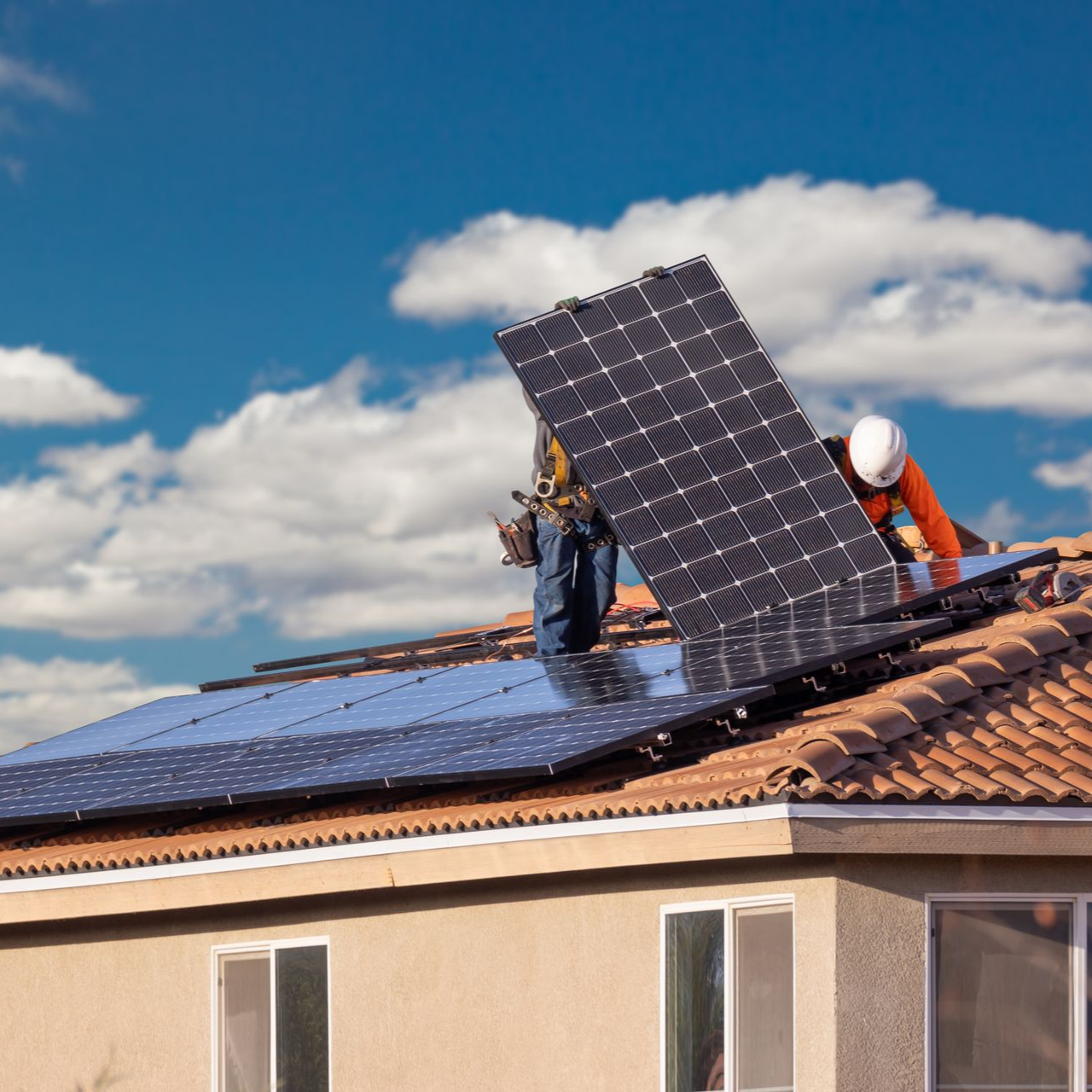 Des ouvriers installent des panneaux solaires sur un toit de tuiles sous un ciel bleu.