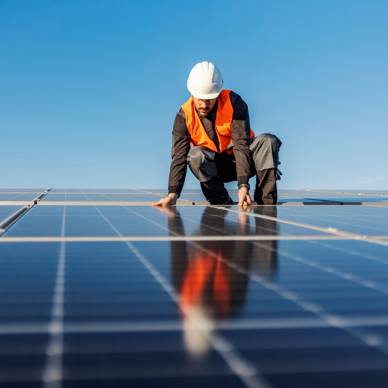 Un ouvrier portant un casque blanc et un gilet orange inspecte des panneaux solaires.