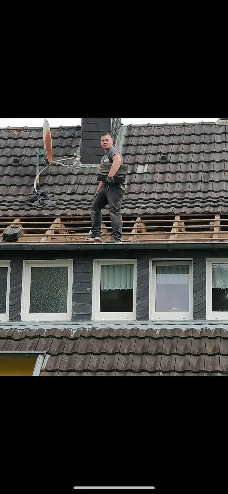 Ein Mann auf einem Dach. Graue Dachziegel, darunter Fenster. Der Mann trägt Hemd und Hose.