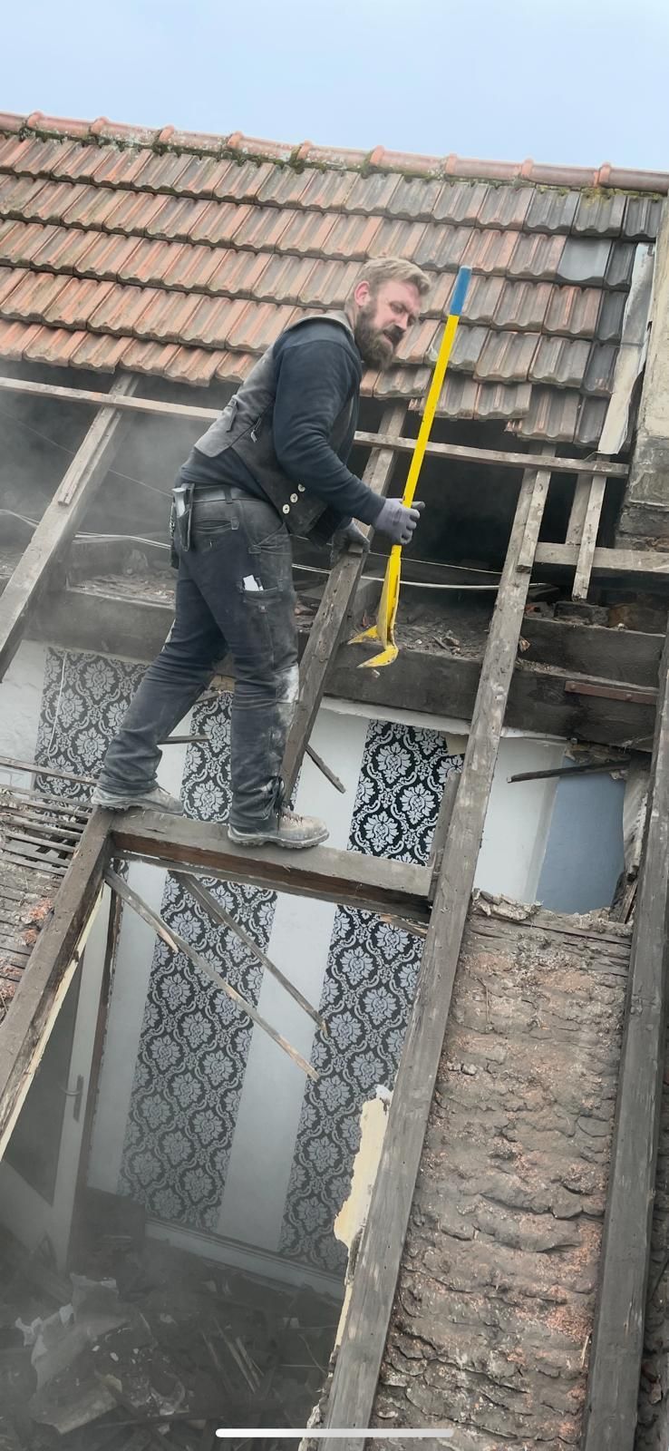 Ein Mann auf einem Dach benutzt ein Werkzeug und entfernt möglicherweise Schindeln oder Bretter. Bewölkter Himmel, freiliegende Holzkonstruktion.