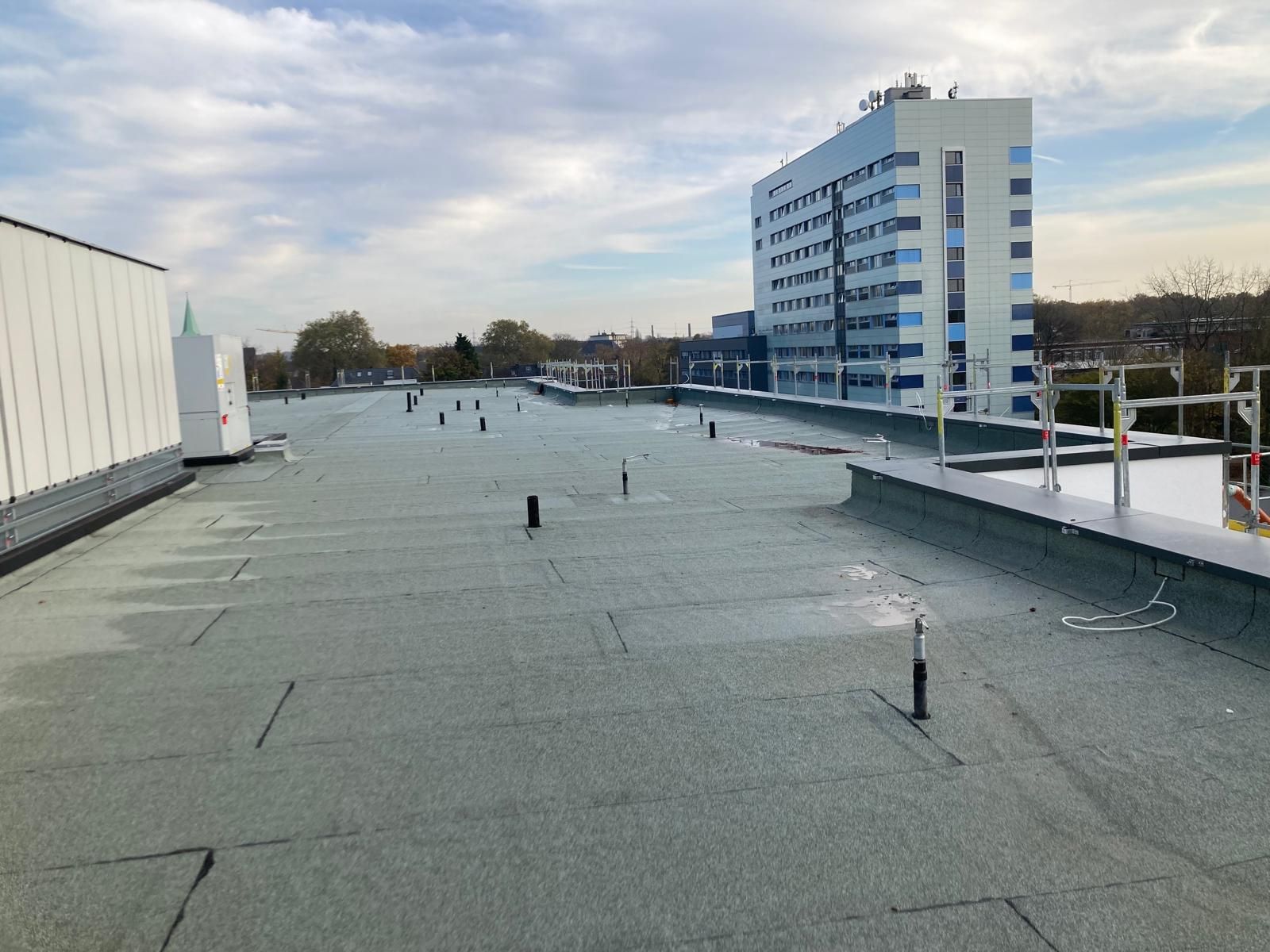 Ein flaches, grünes Dach mit verschiedenen Rohren und einem hohen Gebäude im Hintergrund unter einem bewölkten Himmel.