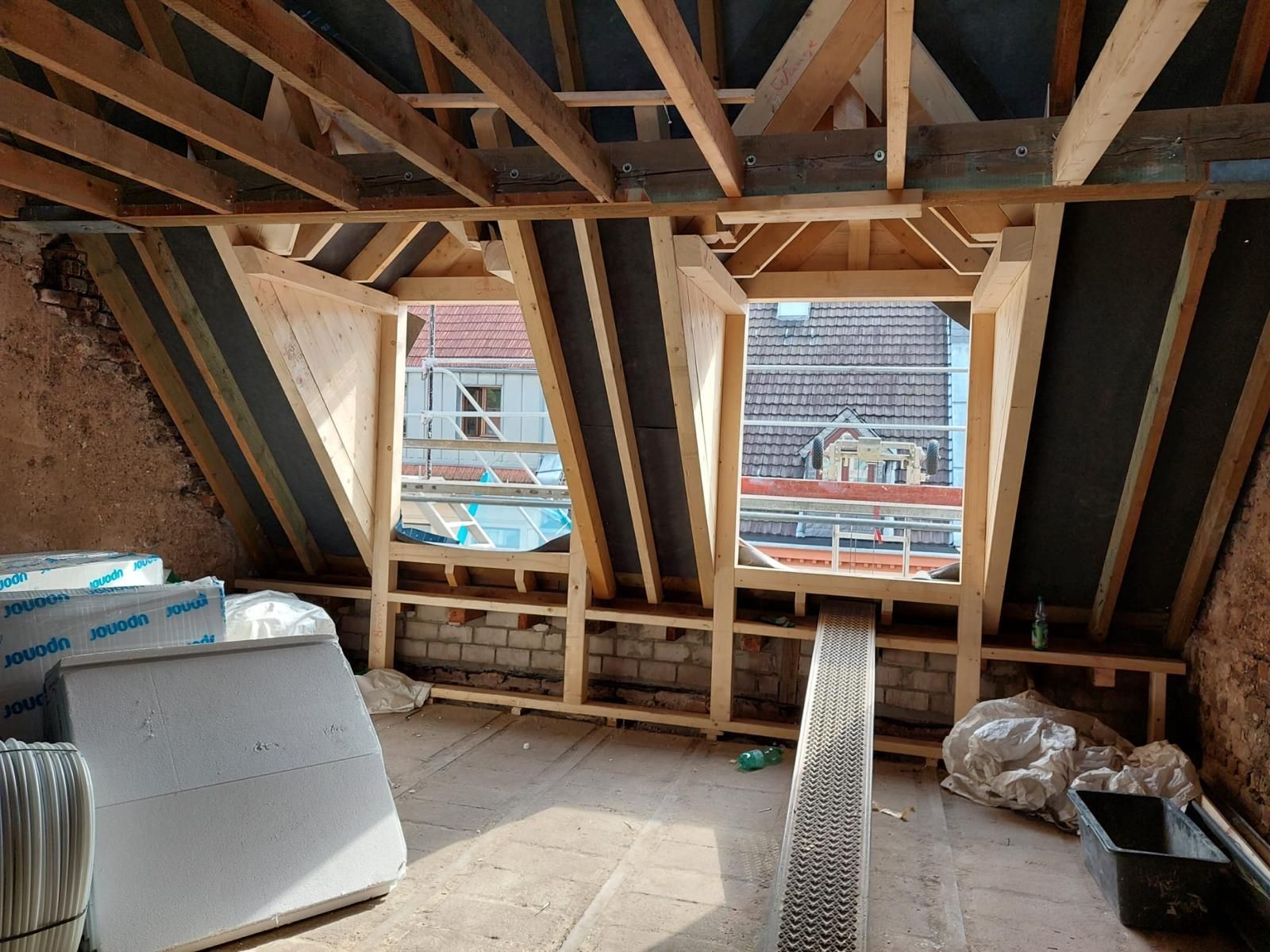 Innenansicht eines im Bau befindlichen Raumes mit zwei Fensterrahmen, freiliegenden Holzbalken und Baumaterialien.