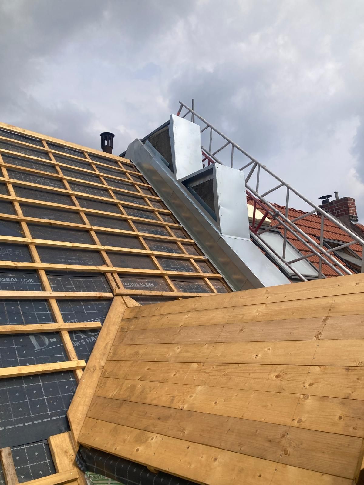 Dach mit unfertigem Holzrahmen, Metalllüftungsöffnungen und einem Sicherheitsgerüst unter einem bewölkten Himmel.