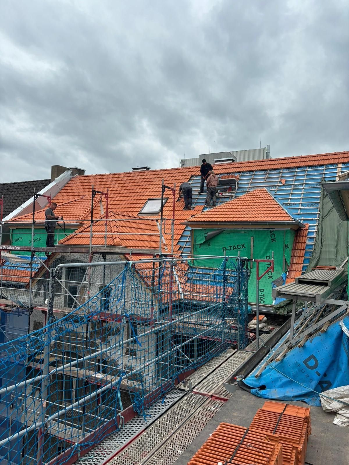 Dachdecker auf einem teilweise gedeckten orangefarbenen Dach verlegen neue Ziegel. Das Gebäude ist von einem Gerüst umgeben. Bewölkter Himmel.