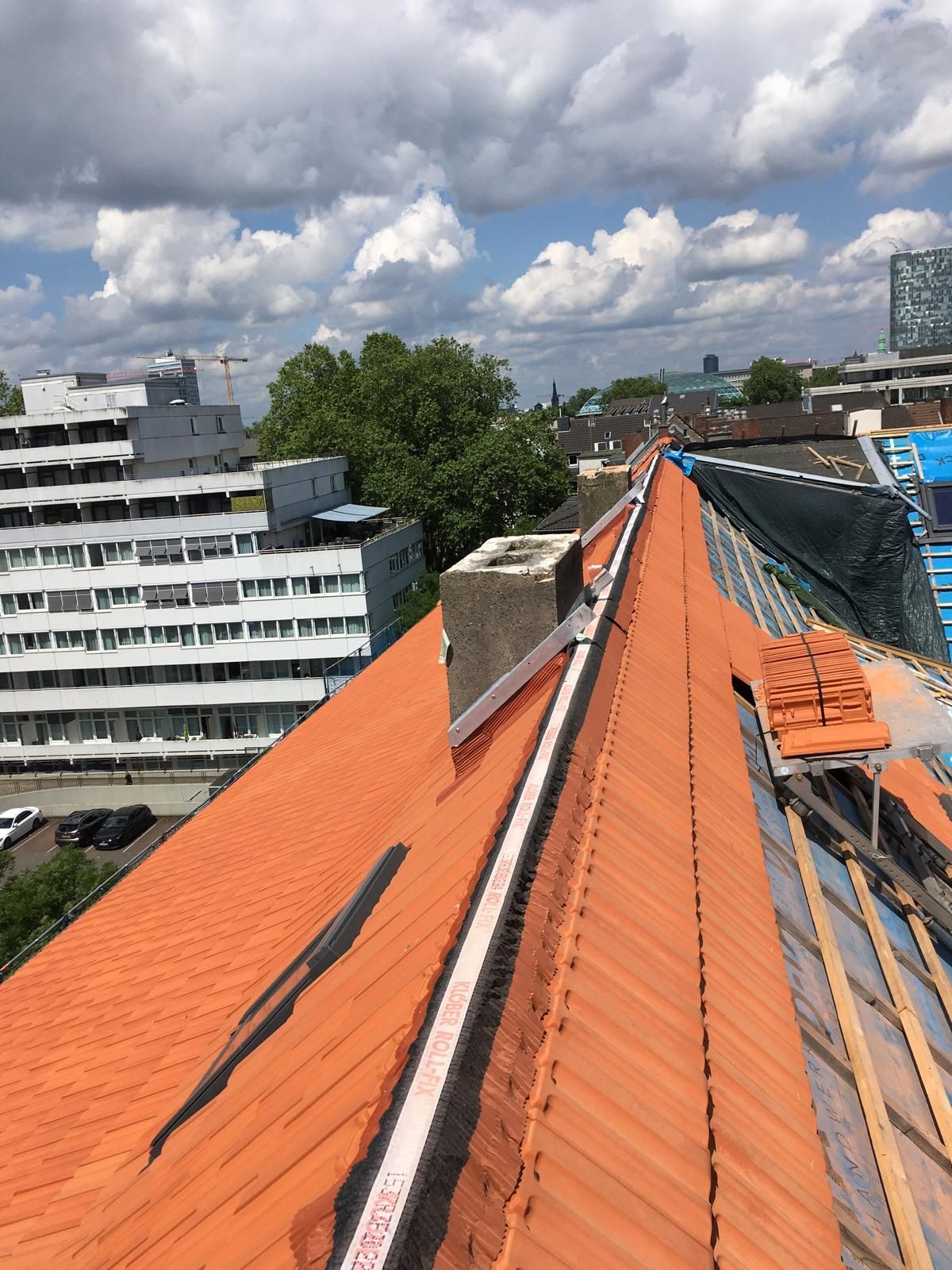 Orangefarbenes Ziegeldach mit Sicherheitsgeländer, Schornstein und städtischen Gebäuden unter einem bewölkten Himmel.