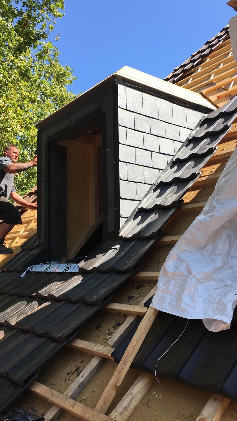 Bau eines Dachfensters auf einem Dach; Mann bei der Arbeit, Schindeln werden angebracht, blauer Himmel.