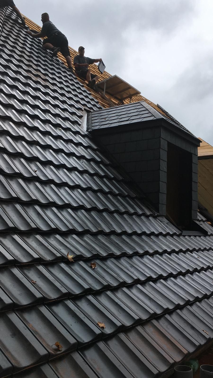 Zwei Dachdecker arbeiten an einem dunklen Ziegeldach in der Nähe eines Dachfensters. Bewölkter Himmel.