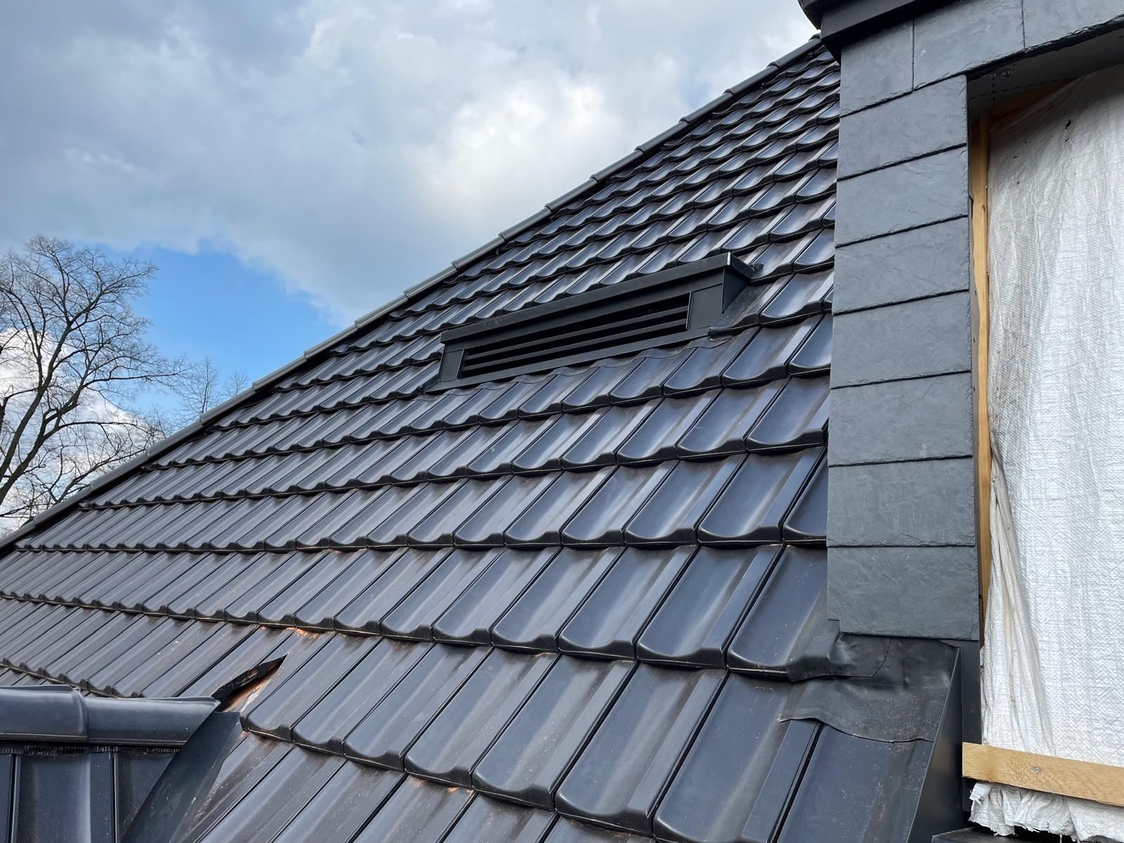 Graues Ziegeldach mit Entlüftung und dunkler Verzierung vor bewölktem Himmel.