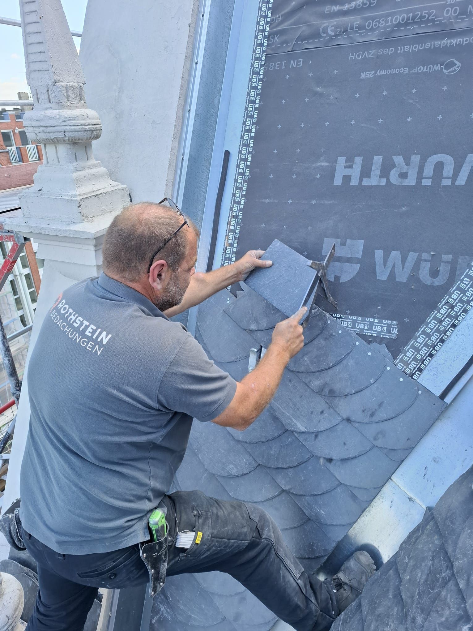 Dachdecker installiert dunkle Schieferziegel auf einem Dach und verwendet dazu Werkzeuge. Graues Hemd, blauer Himmel.