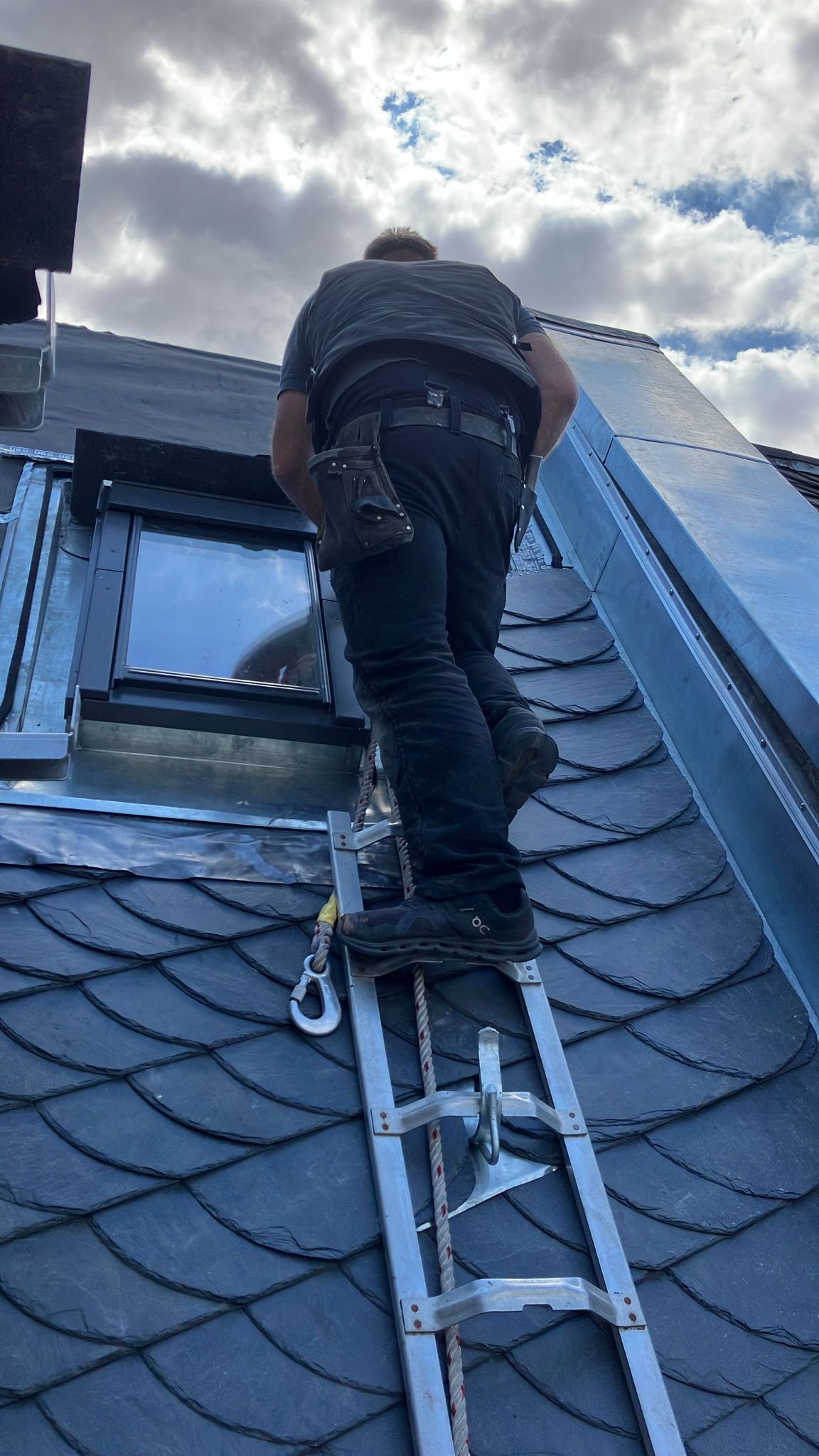 Mann auf Leiter auf Dach, Himmel darüber. Schwarze Kleidung, silberne Leiter und Dach.