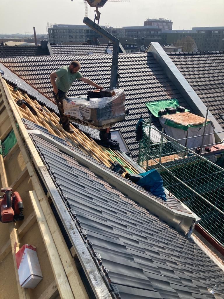 Dachdecker auf einem Schrägdach, der eine Ladung Ziegel mit einem Kran anhebt.