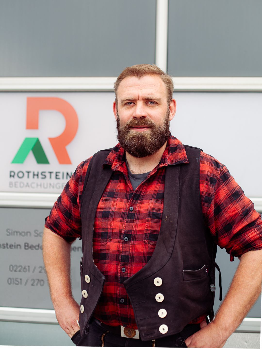 Mann mit Bart und Arbeitskleidung steht vor einer Backsteinmauer.