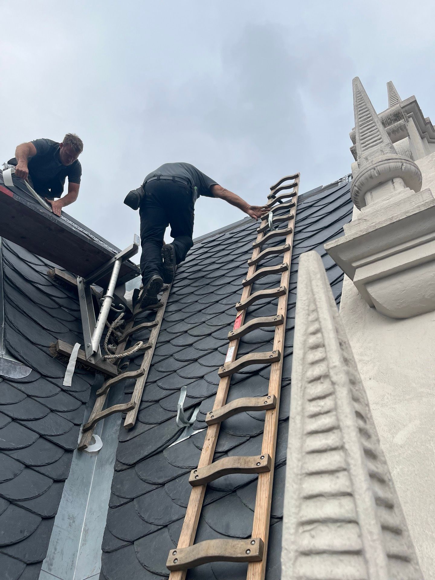 Zwei Arbeiter auf einem dunklen Schieferdach; einer steigt eine Leiter hinauf, der andere in der Nähe eines Gerüsts. Bewölkter Himmel.