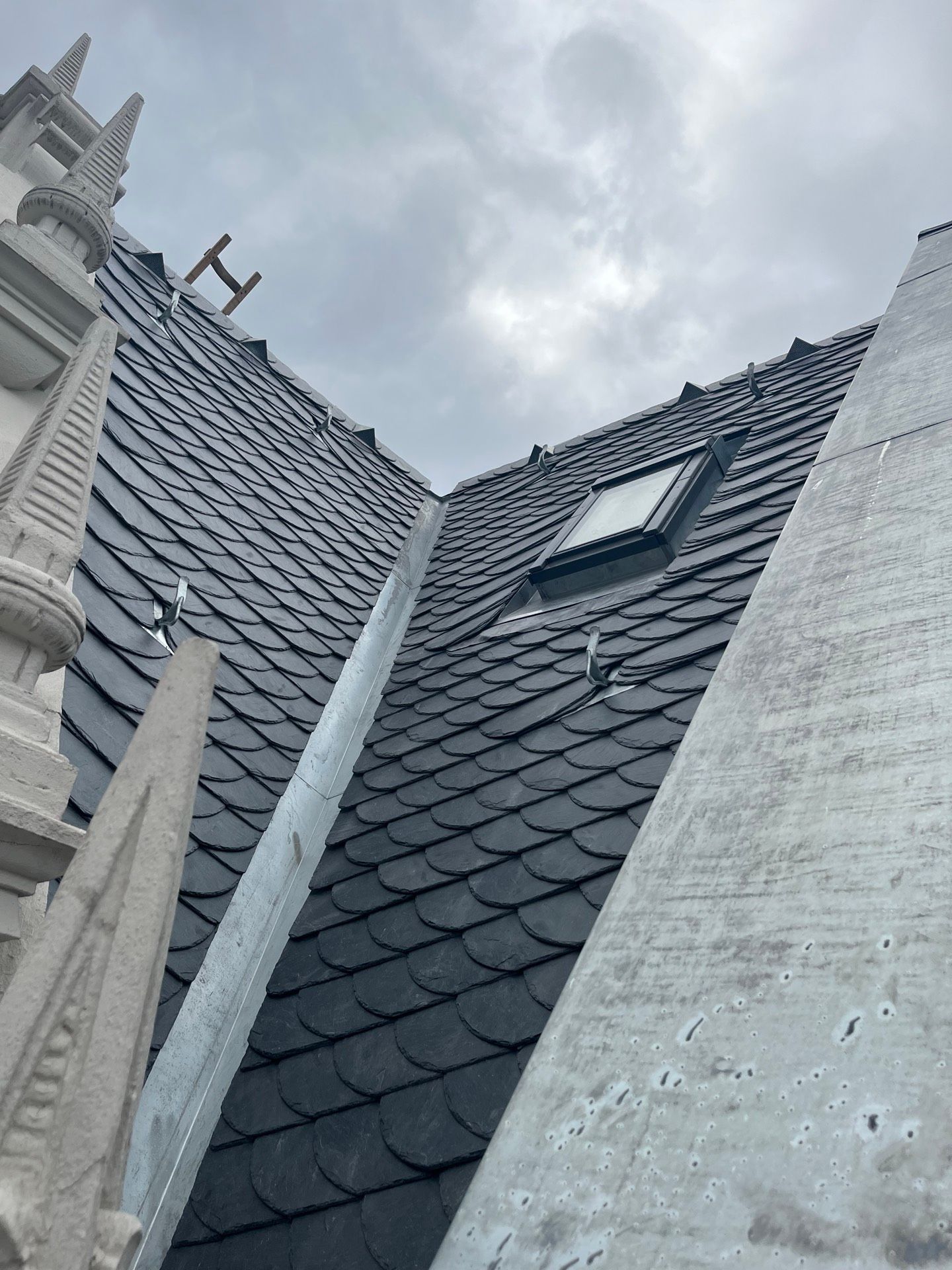 Dunkles Schieferdach mit Oberlicht vor bewölktem Himmel. Metallische Verblendung verläuft in der Mitte.